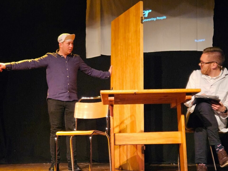 En mann holder en dør midt på scenen og peker til siden. En mann sitter på en stol.