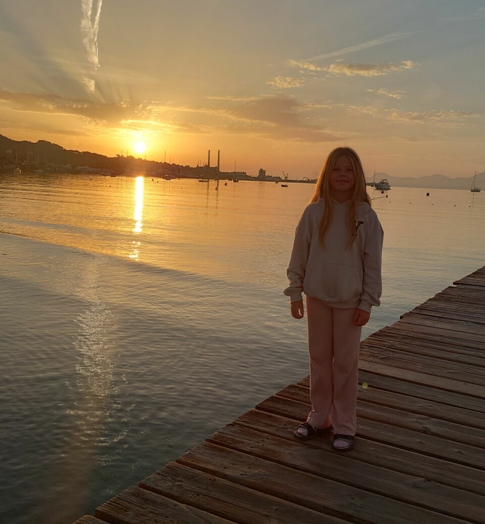 En ung jente står på en brygge ved sjøen i solnedgang