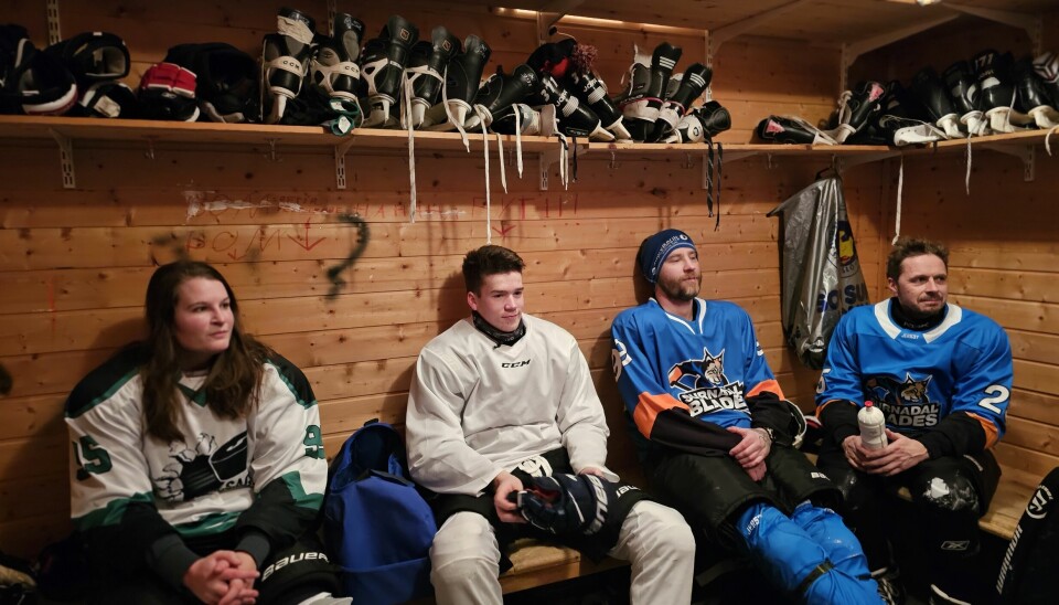 Hockeyspillere fra begge lag sitter på benken i hvilebua. på veggen over dem er det ei hylle fylt med hockeyskøyter.