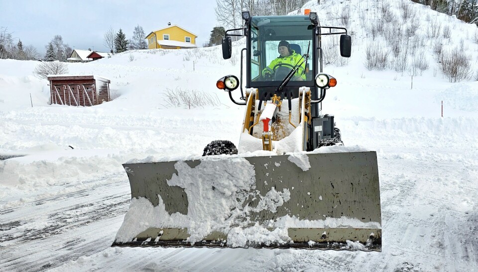 Mann i gul arbeidsjakke sitter i en liten shovel med plog mens veien og parkeringsplassen er delvis ryddet for snø.