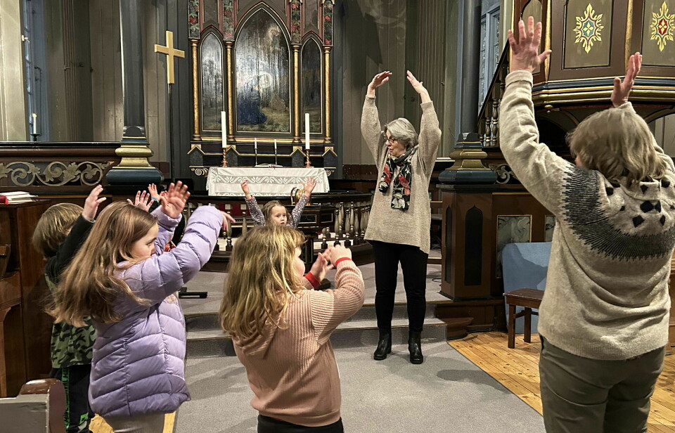 Barn og voksne som som står i en ring inne i ei kirke, og gjør bevegelser med hendene til sang.