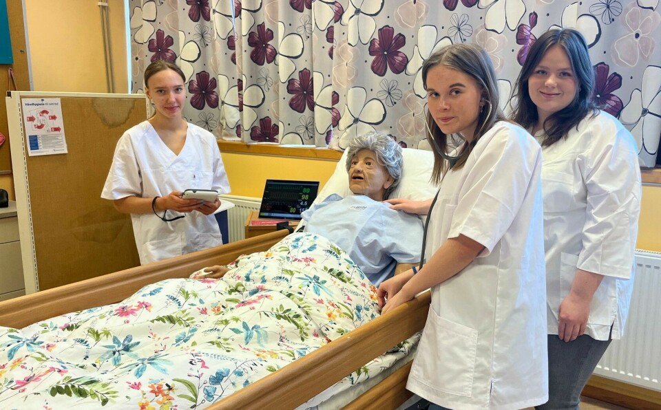 Tre unge jenter i helsearbeideuniform rundt ei seng med ei simulasjonsdukke som forestiller en eldre person.