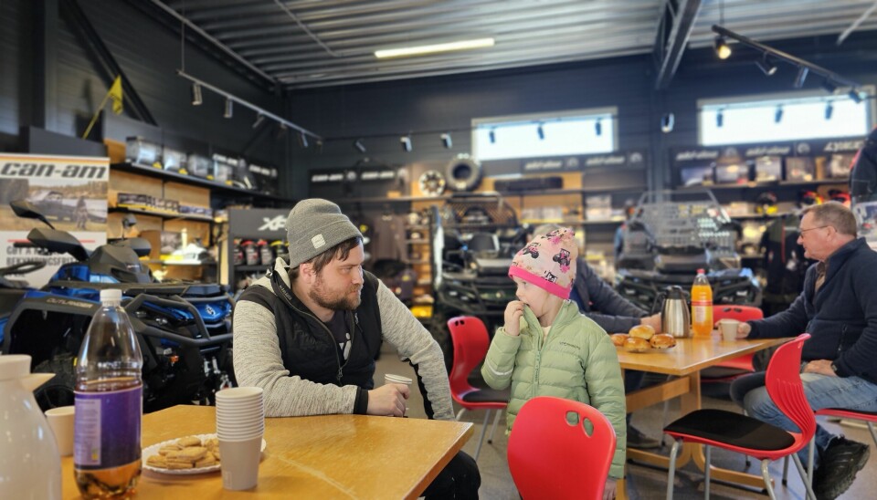 To bord i butikken med kaffedrikkende gjester. I forgrunnen en far med en datter som har rosa traktorlue.
