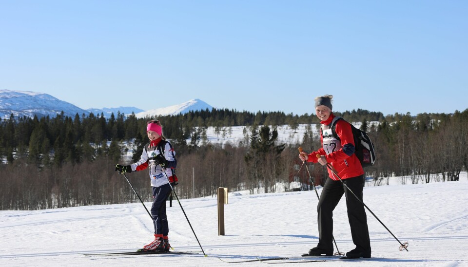 Jente i Rindal IL-drakt og dame i rød skijakke passerer smilende på langrennski. Honnstadknyken i bakgrunnen