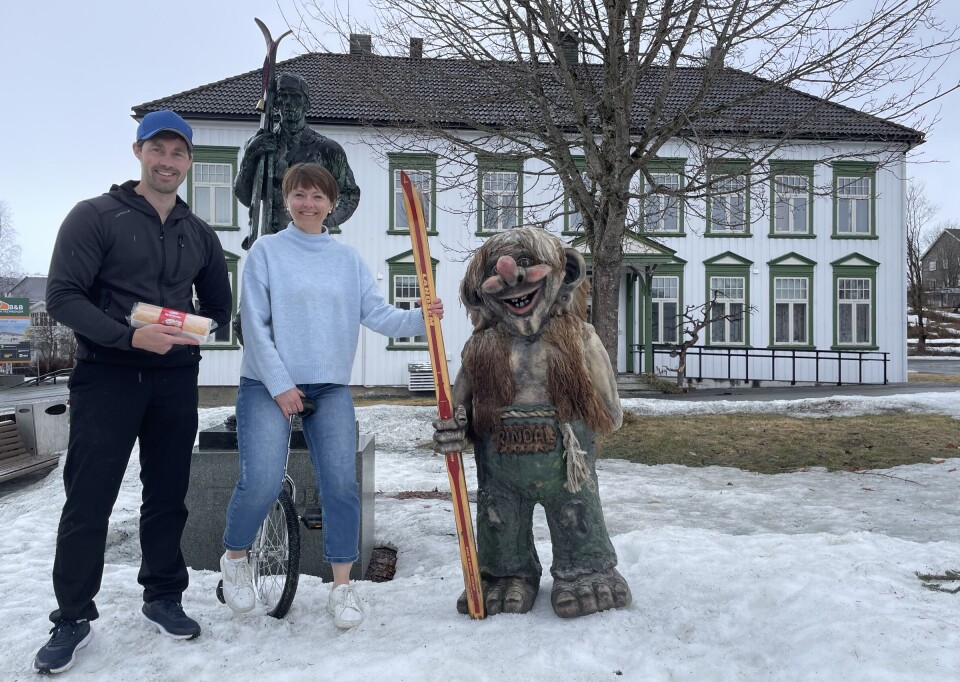 Rullekakebakekonkurransedommer Stian Kooyman og ordfører Mildrid Kattem Aune gleder seg til rullefestivalen. Mann holder en rullekake, dame sitter på enhjulssykkel, og statue av troll som holder ski foran en statue av en mann som holder ski og et hus.