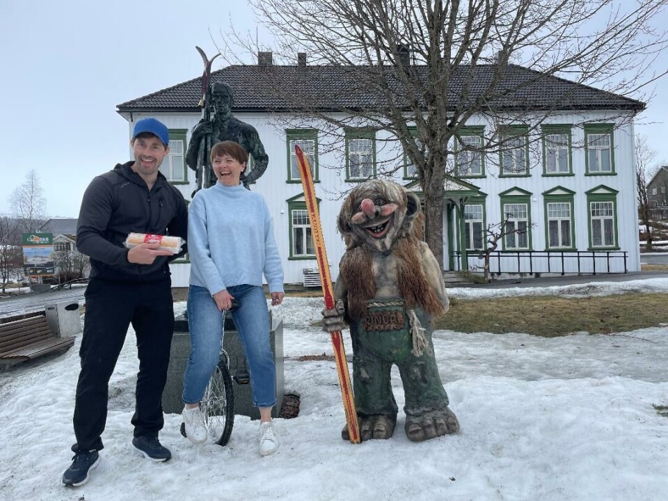 Mann holder en rullekake, dame sitter på enhjulssykkel, og statue av troll som holder ski foran en statue av en mann som holder ski og et hus.