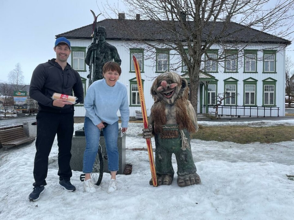 Mann holder en rullekake, dame sitter på enhjulssykkel, og statue av troll som holder ski foran en statue av en mann som holder ski og et hus.