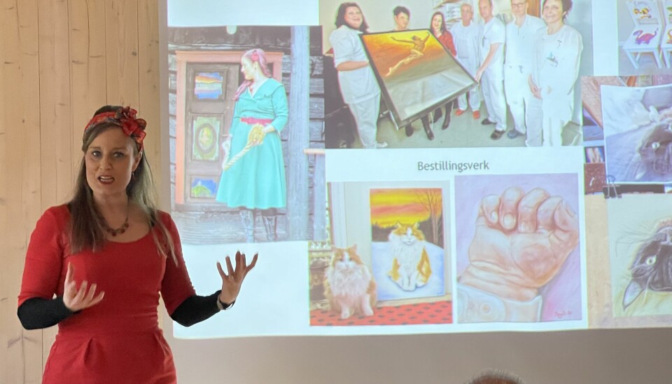 En dame i rød kjole og mørkt hår står foran en skjerm med ulike bilder. Ett med seg selv i blå kjole utenfor en dør, en med to katter, en med en barnehånd, og et bilder hvor sykepleiere viser frem et bilde