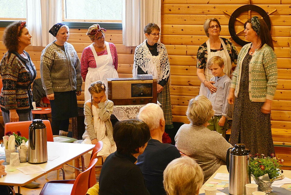 Damer i gamle klær synger med publikum sittende rundt bord i salen