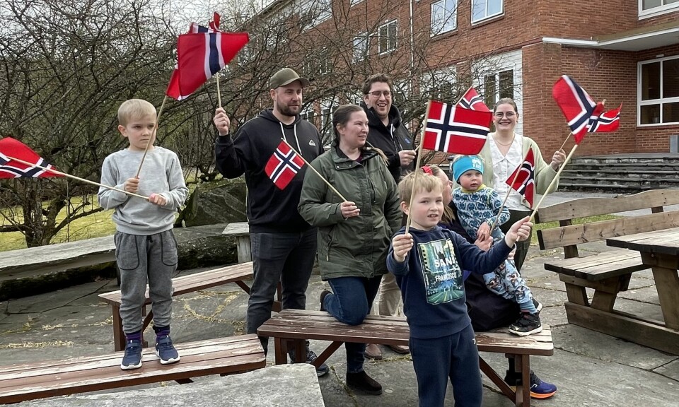 3 barn og 4 voksne med norske flagg. Står i en klynge ved noen benker foran en murbygning. Trær uten blad i bakgrunnen