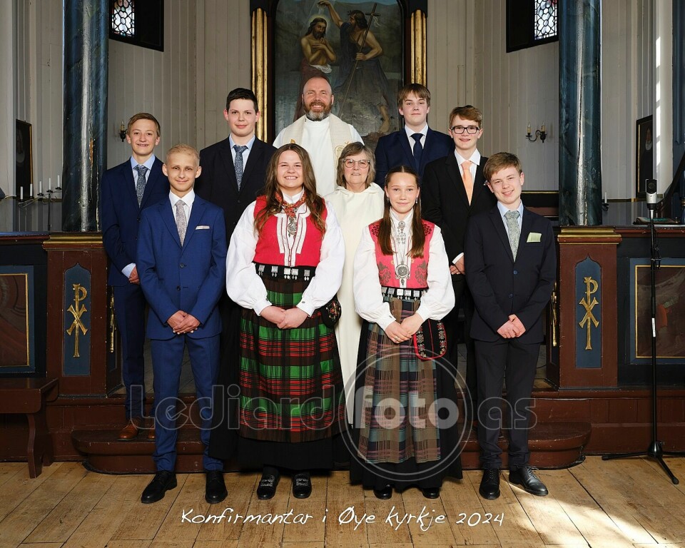 Åtte konfirmantar, ei dame i kvit kappe og ei prest, oppstilte for fotografering framme i koret i kyrkja.