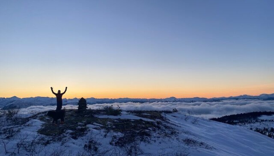 Følelsen av mestring er stor når man er kommet på toppen. Her også over skyene som ligger over fjorden. En liten person står på toppe av et fjell med hendene opp over hodet. Bakgrunnen ser man flere fjell og lavt skydekke over fjorden