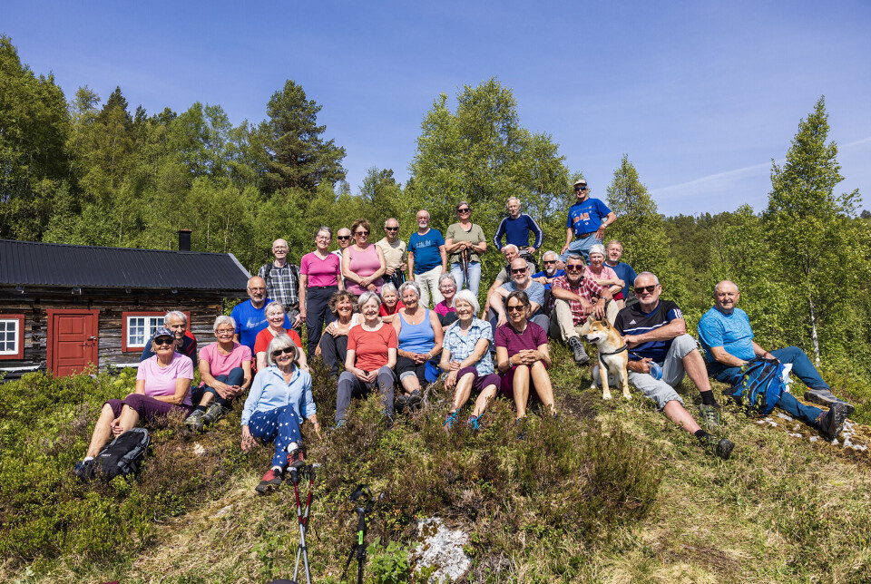 30 spreke pensjonistar framom Oppistusetra Solem. Mange sommerkledde turgåere sitter eller står på en haug foran et gammelt seterhus.
