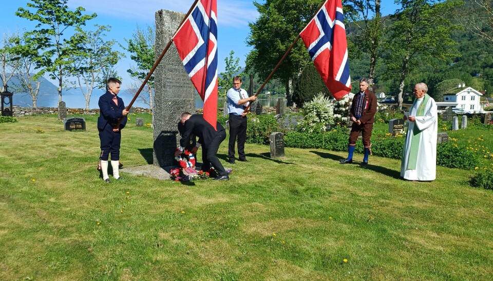 Ordfører Hugo Pedersen legger ned blomster på graven til Halvor Røen. ørdfører bøyer seg og legger blomster på stor grav, to menn med norske flagg på hver side av graven