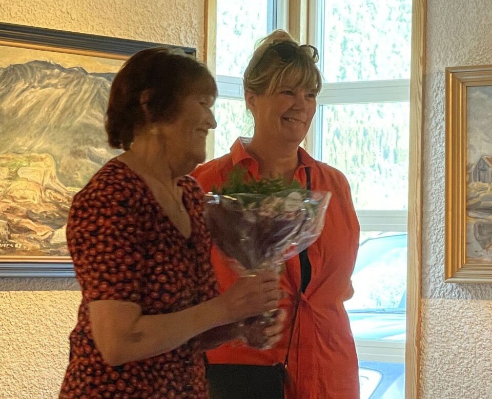 Inger Marie Gjøra og Lilli Husby forteller om Lars Gjøra. To damer i rød kjole, den ene med en blomsterbukett i hånden. Maleri i bagrunnen