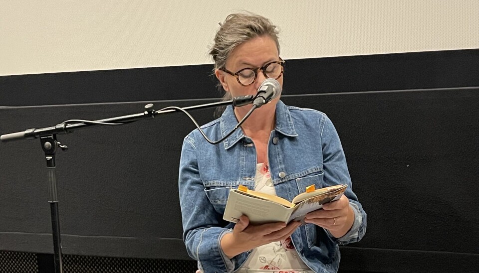Dordi Strøm leser høyt fra sin siste roman Odel En dame sitter å leser en bok. Microfon foran seg. Hun har på hvit kjøle med roser og dongrijakke