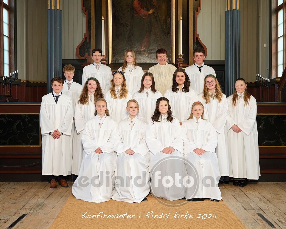 Konfirmantene i Rindal kirke 16 konfirmanter i hvite kapper og en prest oppstilt til fotografering i koret i kirka.