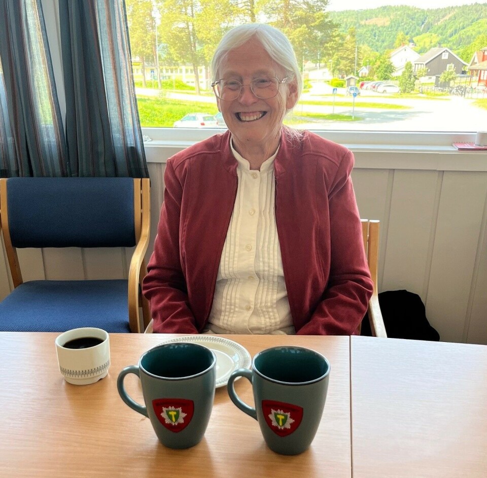 Linda fikk to krus av brannvesenet. Disse kan hun ha på hytta, om hun vil. Ei smilende dame som sitter ved et bord med en kaffekopp og to krus foran seg.