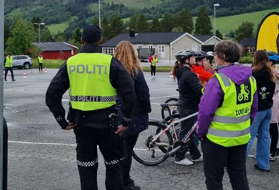 En politibetjent i uniform og gul varselvest, ei dame i gul varselvest med NAG-logo, og flere barn på sykler. NAF-flagg og flere personer i gule vester i bakgrunnen.