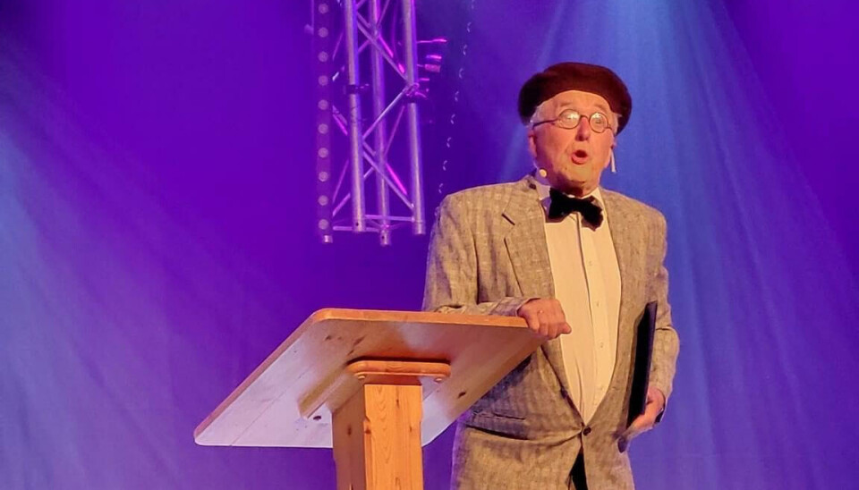 Øyvind Holte som professor Hansen, en fornøyelig type En eldre mann står ved en talerstol. Grå dress og alpelue