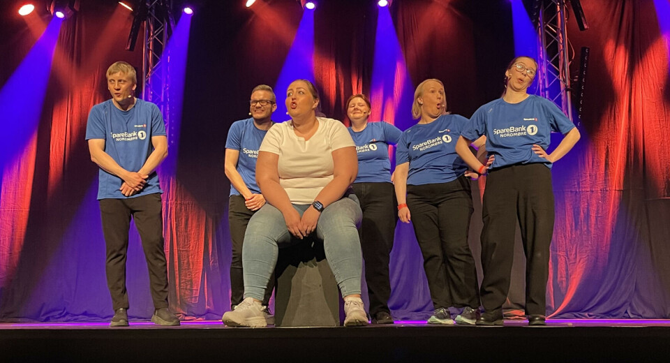 En sterk opptreden av gjengen fra Svarrabærje teaterlag En person i dongribukse og hvit t-skjorte sitter på en scene. bak henne står 5 personer med svart bukse og blåe t-skjorter. Blått og rødt scenelys i bakgrunnen