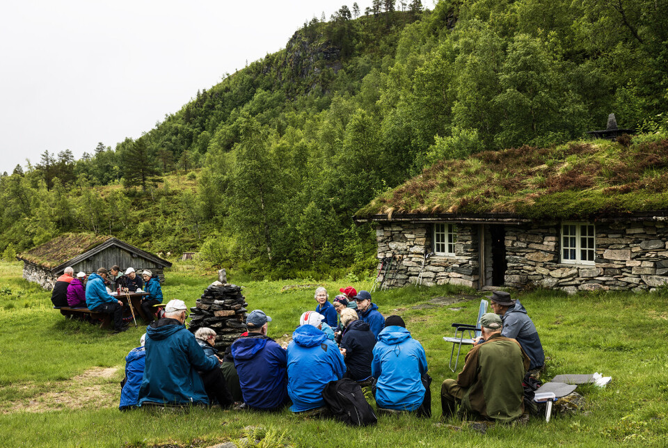 Matpause på setervollen på Ranessetra. Mange turkledde mennesker som sitter og spiser nista si på et gammelt setertun, med to gamle seterbyginger av stein.