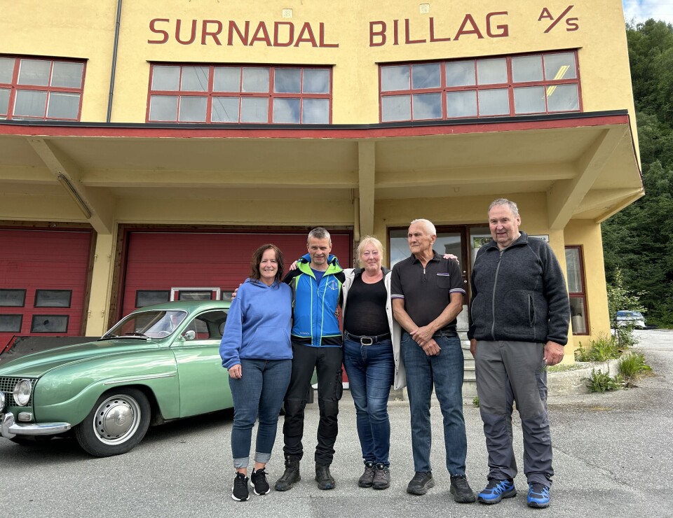 Fem styremedlemmer med ulik fartstid. Fem personer foran en murbyging med påskrift Surnadal billag A/S, og en veteranbil bak.