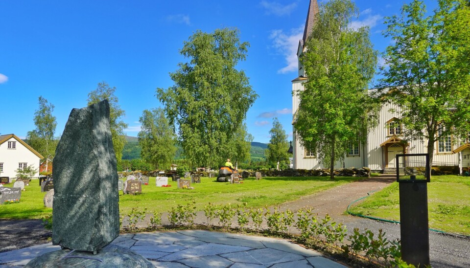 I jubileumsåret til Rindal Kirke blir det åpning av minnelund og utendørs fotoutstilling. Kirka i bakgrunnen, en mann sitter på plenklipper og deler av minnelunden og minnesten i front.