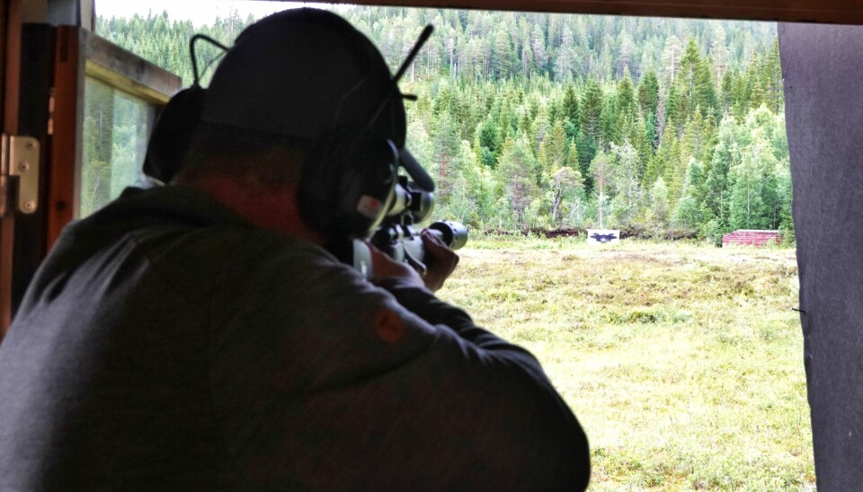 En stor skytter med geværet i posisjon sees bakfra og vi kan skimte plata med den bevegelige 'elgen' i bakgrunnen mot skogen.