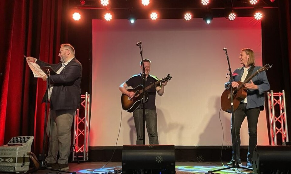 Odd Arne Halaas fikk to dialektplakater som takk for jobben på Optimistkvelden. En plakat med hans egen Eidedialekt, og en med Rindalsdialekt, så klart. Han lurte litt på ordet pøngelh^k, hva det kan bety. Kunne det være en man ikke vil møte ute kl 3 om natta? Han fikk vel bekrefta at det i alle fall ikke er helt feil. En mann på en scene som leser fra en plakat, to menn med gitar ser på han og ler.
