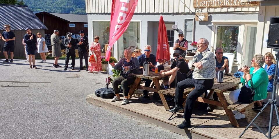 Bernt Bøe kåserer om historien rundt butikken, og las opp eget dikt om karene som satt på klagemuren. Mange folk som sitter utenfor en Coop Marked-butikk i sommervær. En mann sitter og snakker i mikrofon.