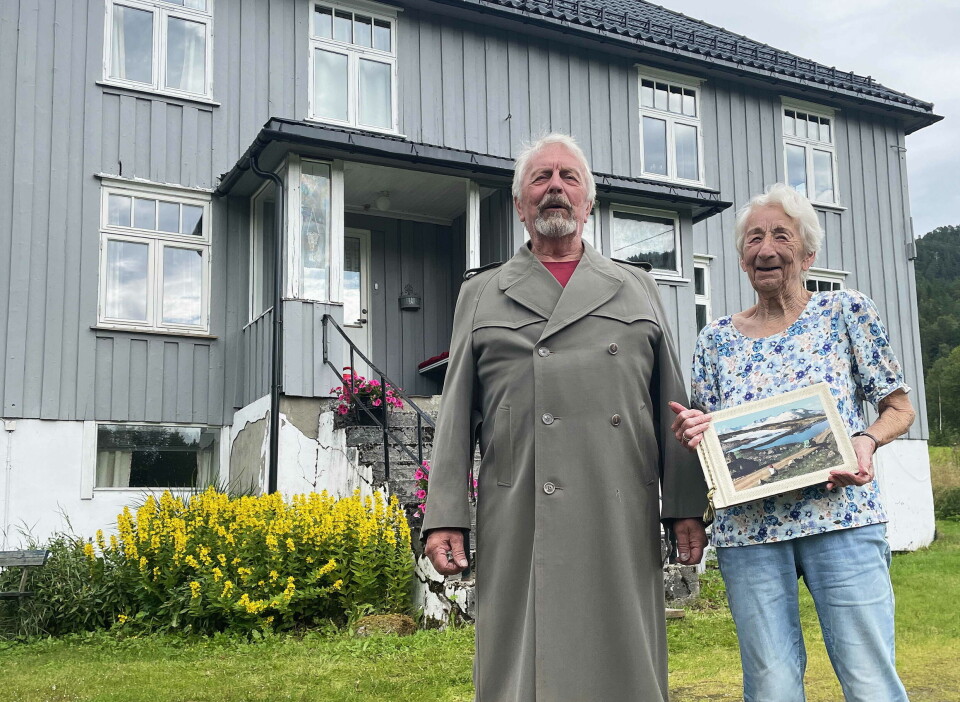 Søskenbarna Geir og Elise Settemsdal framfor «verkstedet» som husa konfeksjonsfabrikk, butikkar og posthus. Geir iført ekte kappe frå fabrikken! En mann og ei dame foran et gammel hus.