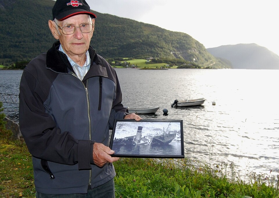 - Ved holmen bak meg forliste rutebåten i mars 1944! Asbjørn var passasjer og viser her foto av havaristen. Mann står ved fjorden med et bilde i hånda