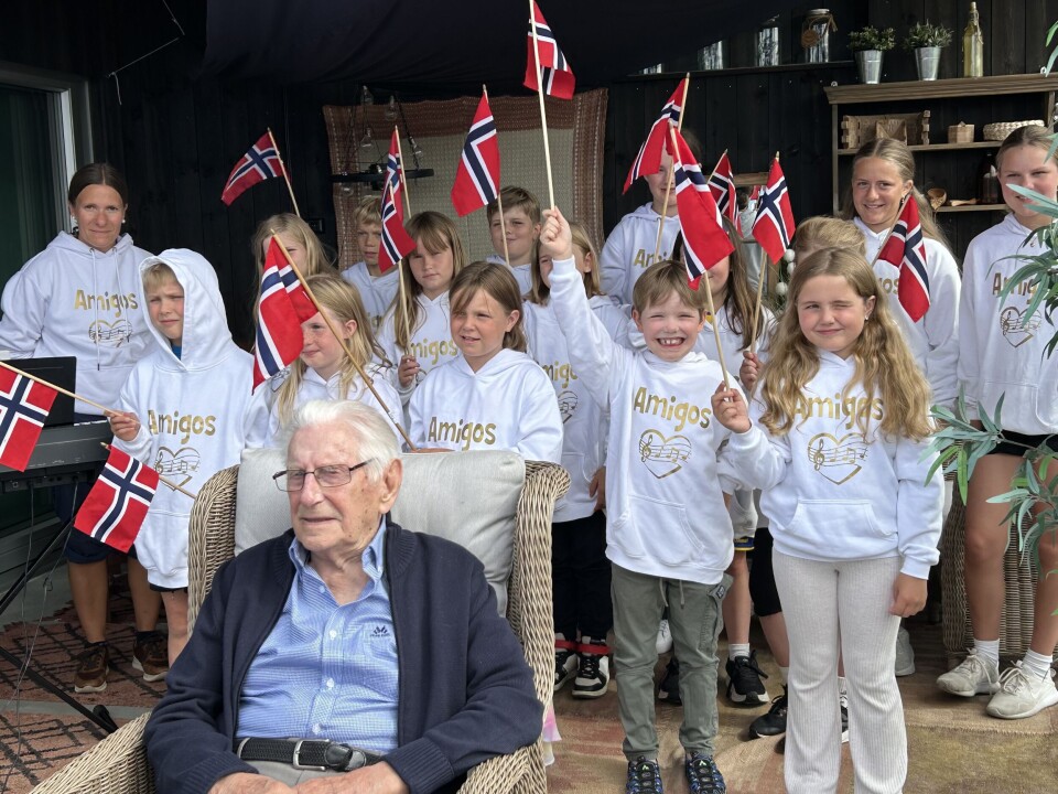 En sangglad gjeng og en overraska jubilant! En mann sitter i en stol og bak han står barn med flagg