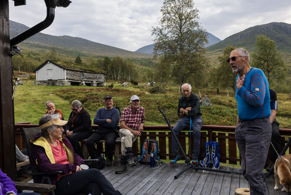 Oddvar Kvande fortel svært interessant om setringa på Valsetsetra. En mann som står foran ei gruppe turkledde mennesker og snakker. De sitter på en veranda på ei fjellhytte, med et seterfjøs i bakgrunnen.