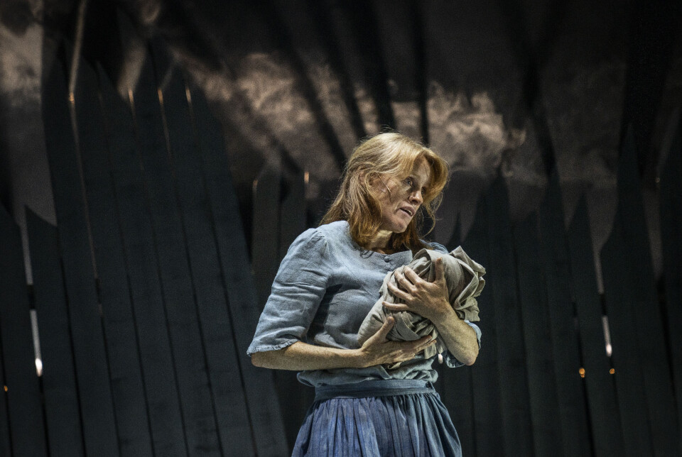 Kjersti Sandal i Jordmor på jorda. Ei dame som står på en teaterscene, med noe som ser ut som en bylt med et nyfødt barn i hendene.