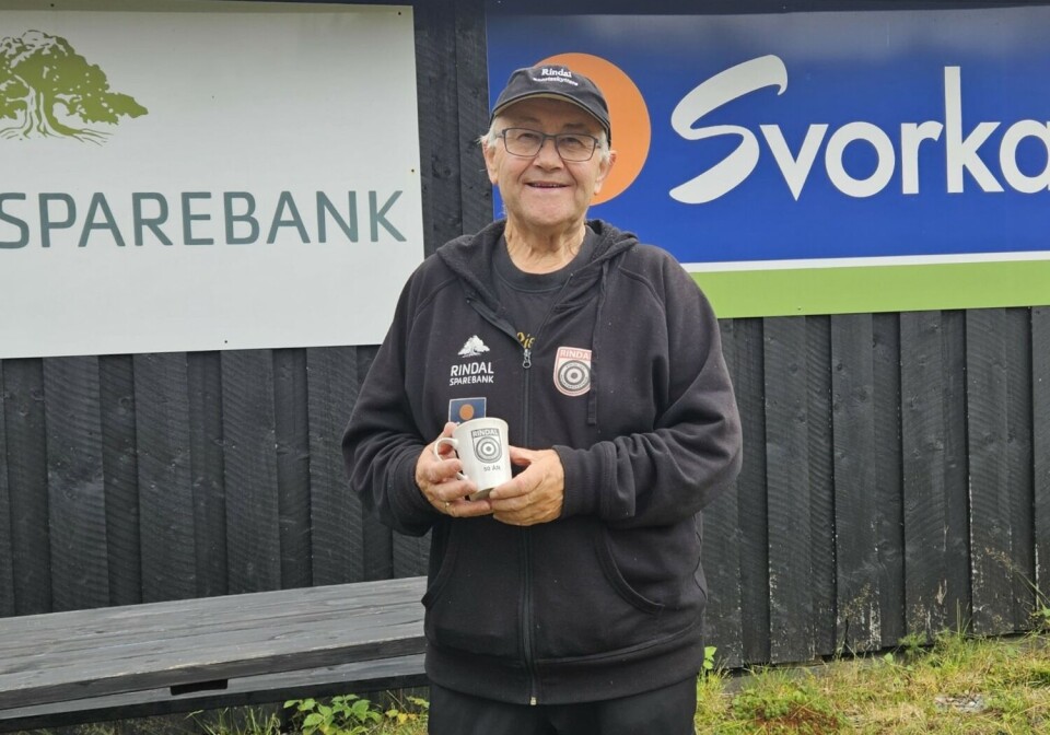 Rindal Sportsskytteres leder fra stiltelsesåret 1974 frem til i dag, Idar Moe. Her med jubileumskruset Mann med krus foran reklame på skytebanevegg