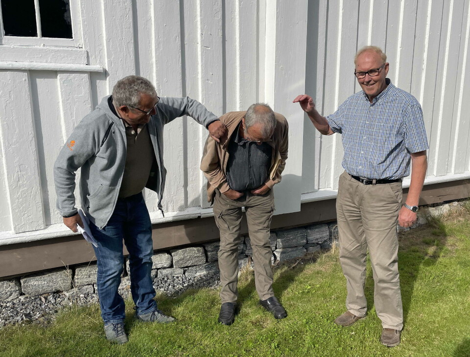 Attmed kyrkjeveggen skal Einar Oterholm (i midta) settast i sjøllaga gapestokk. Lensmann Vatten (t.v.) og jubileumsleiar Indreiten ser til at det går rett for seg. Tre mann står foran kirkevegg. To står med bøyd rygg.