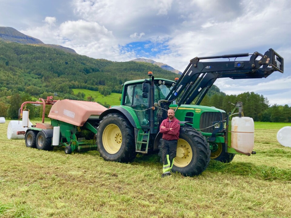 Håvard Halset er godt i gang med 4. slåtten, og er fornøyd med årets avlinger. Stor traktor og kombipresse bak. Mann står med armene i kryss foran traktoren.