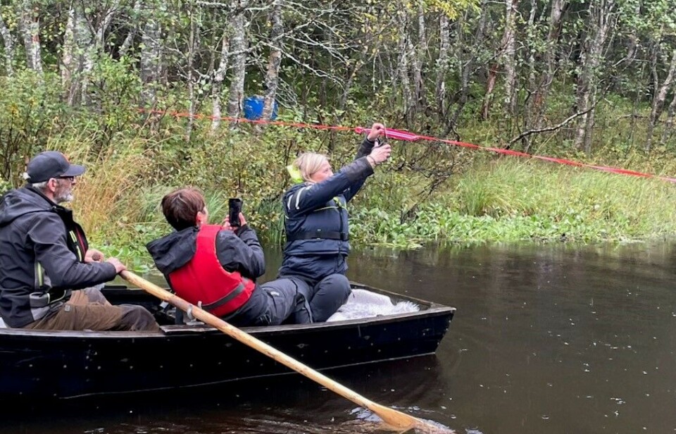 Snorklipping. Tre personer sitter i en lite robåt på ei elv. Ei dame som sitter i båten klipper ei rød snor som er spent over elva.