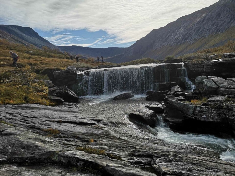 Fossen i utløpet av Vassdalsvatnet. En liten foss i et fjellandskap. Noen turgåere i bakgrunnen.
