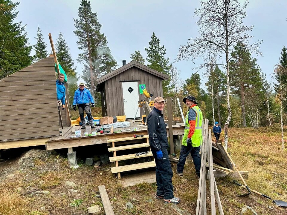 Nå blir det gapahuk ved Haraldhøtta på Tiursåsen. Baard i full sving sammen med dugnadsgjengen mandag kveld. Folk som snekrer på en gapahuk ved en platting og ei lita hytte. Baard med gul snekkervest i front.