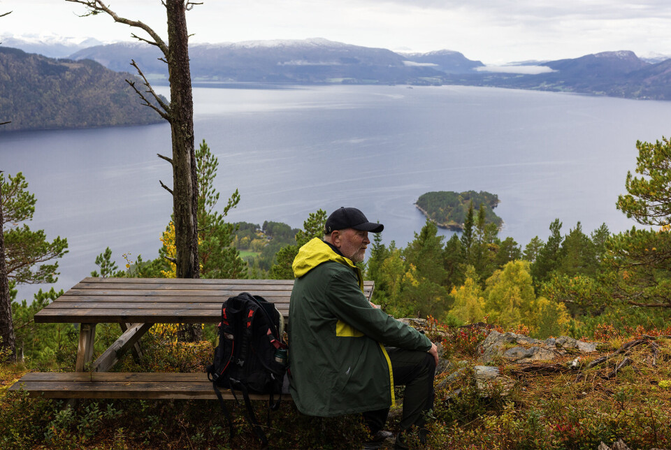 Utsikt mot fjordane. En turkledd mann som sitter på en benk på et utsiktspunkt. Fjord og fjell i bakgrunnen.