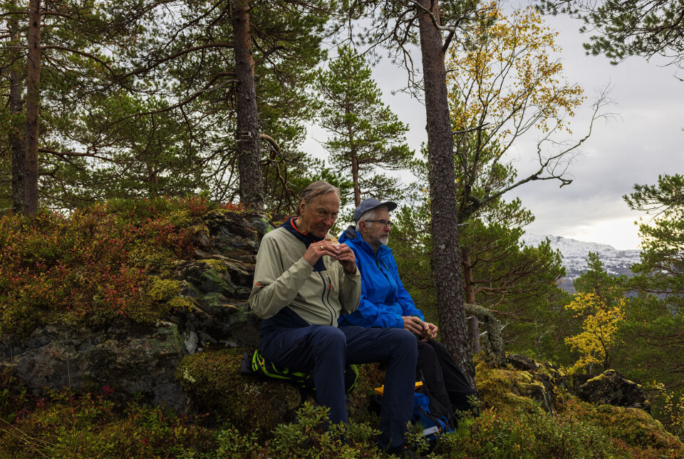 To som nyter nista på tur. To turkledde menn som sitter i skogen og spiser mattpakka si.