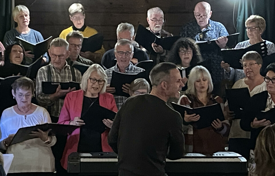 Engasjert dirigent som både spiller piano, synger med og holder alle samlet. En person med ryggen til midt i bildet. Ser til høyre. Deler av et sangkor bak. Både menn og kvinner. Alle med svarte permer