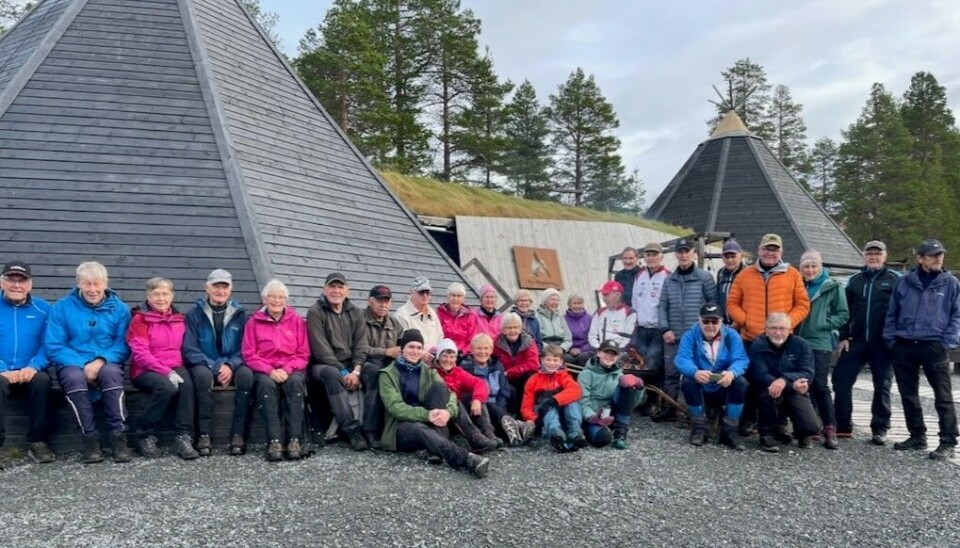 31 turgåere på avslutninga av tirsdagsturen ved Trollheimstunet. Mange turgåere oppstilt for fotografering foran bygninger utformet som lavvoer av tre.