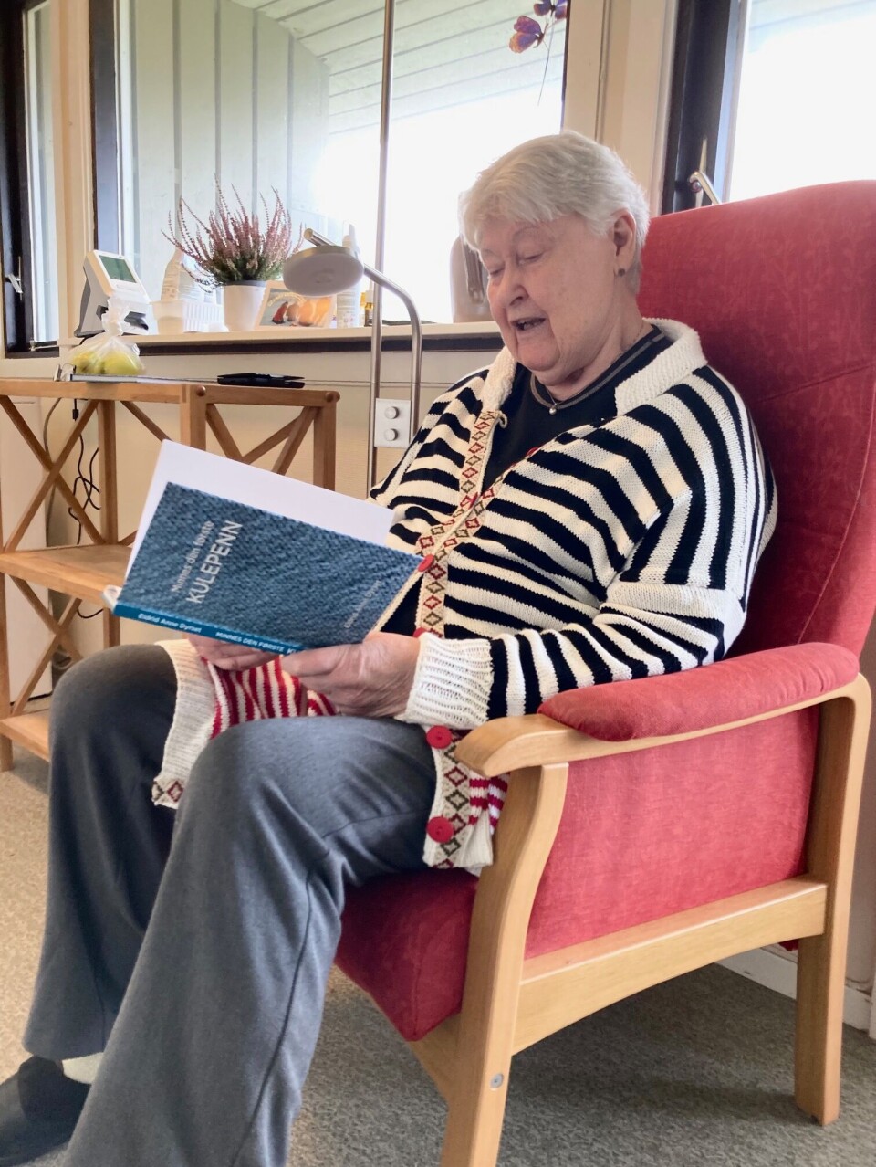Eldrid Anne Dyrset er stolt av å ha gitt ut bok i en alder av 76 år. Eldre dame med stripete genser, bok og rød lenestol