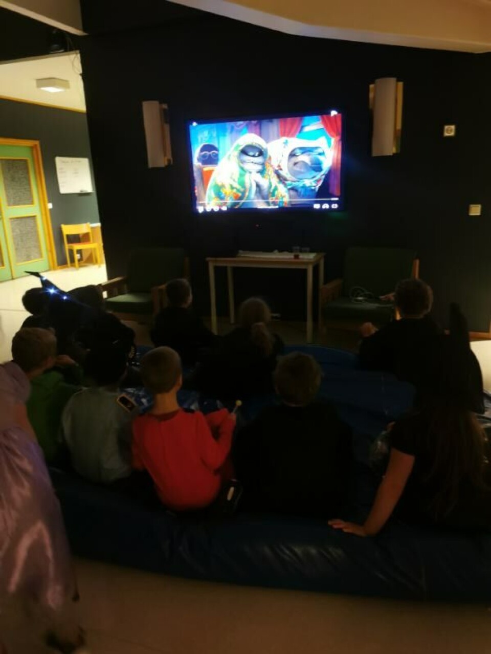 Her er det film i vestibylen. Hotell Transylvania 3. Med mye godteri. Barn på tjukkas som ser på TV.