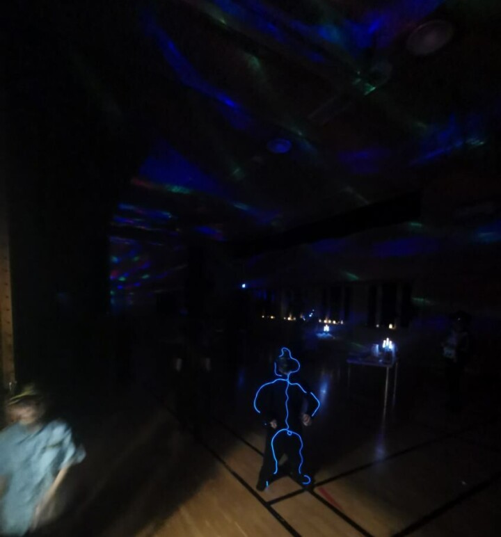 Musikk og dans i gymsalen. Diskolys i taket i en mørk gymsal, en unge med selvlysende drakt i forgrunnen.