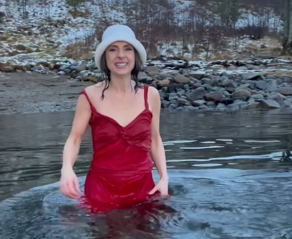 Jenny Klinge under innspeling av julevideo. Ei dame som står i sjøen med rød kjole. Det er snø på marka bak ho.